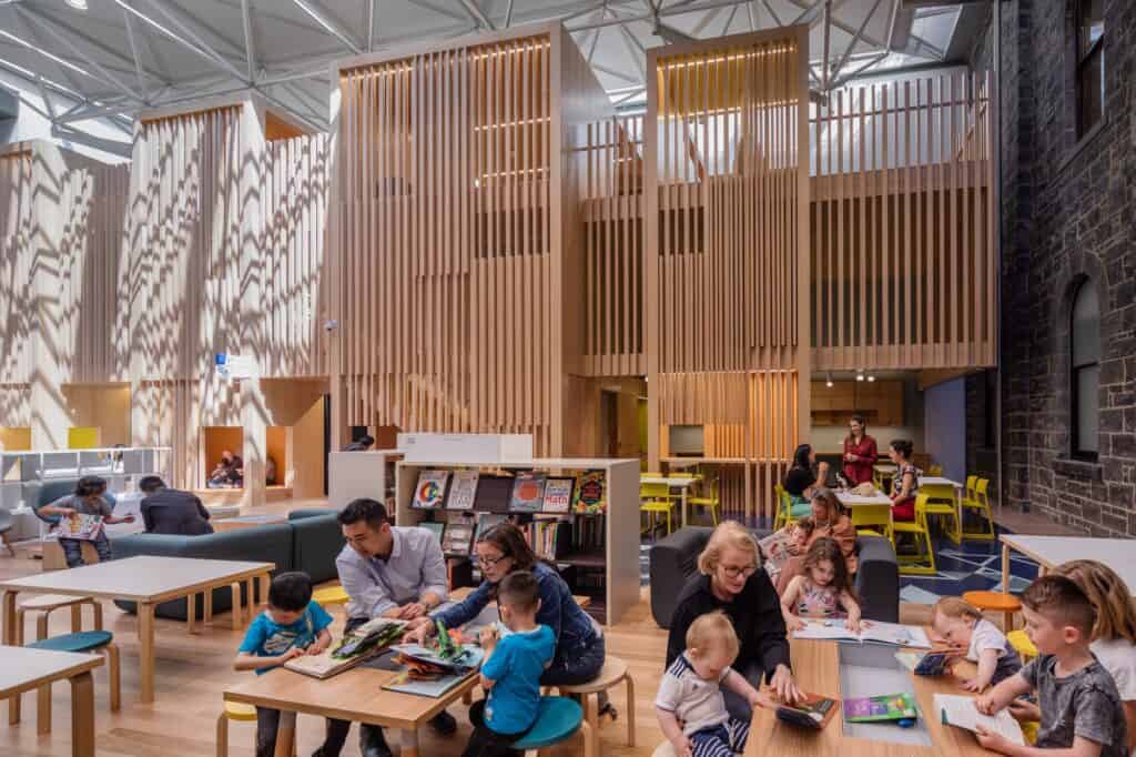 Children and parents reading at low timber tables in the Pauline Gandel Children's Quarter at State Library Victoria, timber batten towers rising under a skylit roof beside an old bluestone wall