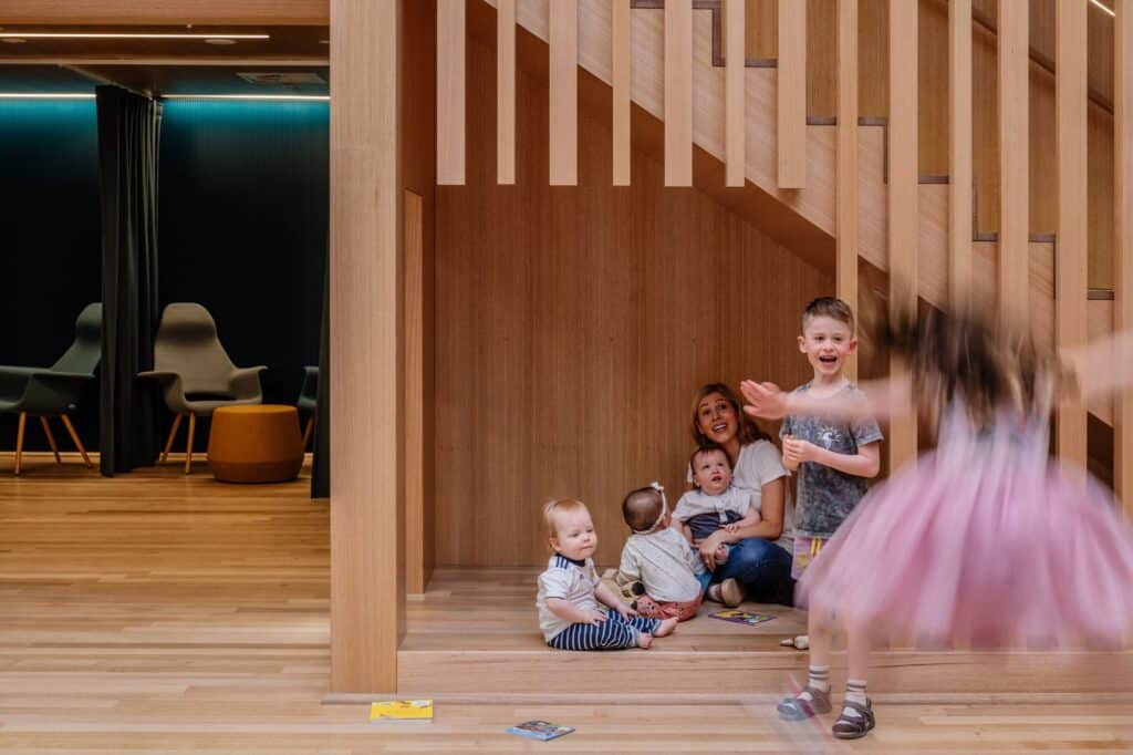 Toddlers, a boy and a mother sitting in a timber nook under the stair of the children's castle at State Library Victoria, a girl in a pink tutu twirling in the foreground