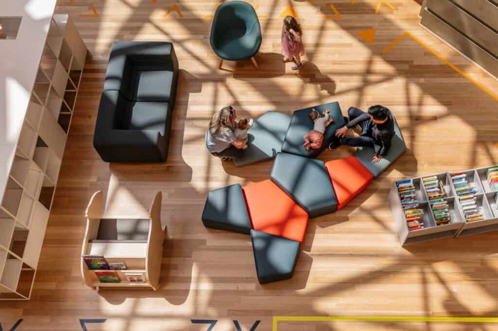 Overhead view of the Children's Quarter at State Library Victoria, children lounging on geometric charcoal and red floor cushions beside a navy sofa, low white bookshelves and dappled light on the timber floor