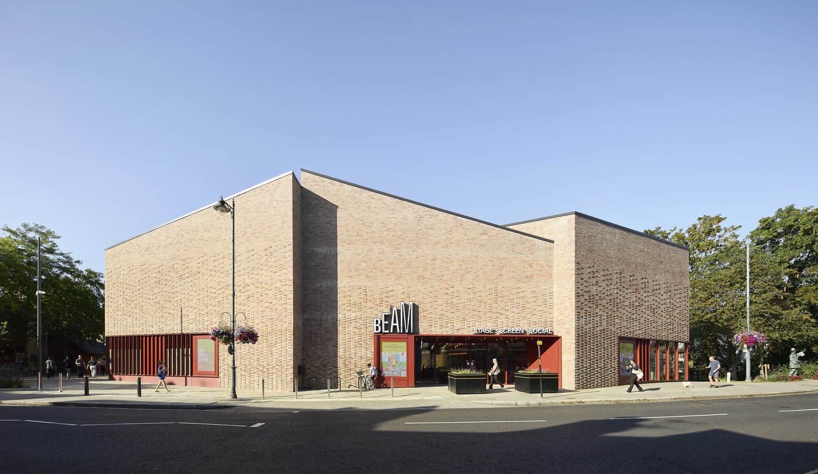The main entrance of BEAM in Hertford, a large angular building clad in buff brick with a red-framed glazed entrance at its base, seen across a quiet street on a clear day