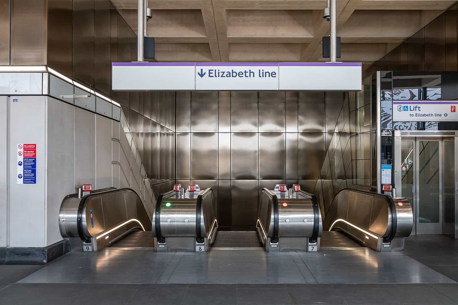 The Elizabeth line entrance at Farringdon station in London, with three escalators descending between brushed stainless steel walls below a purple Elizabeth line sign
