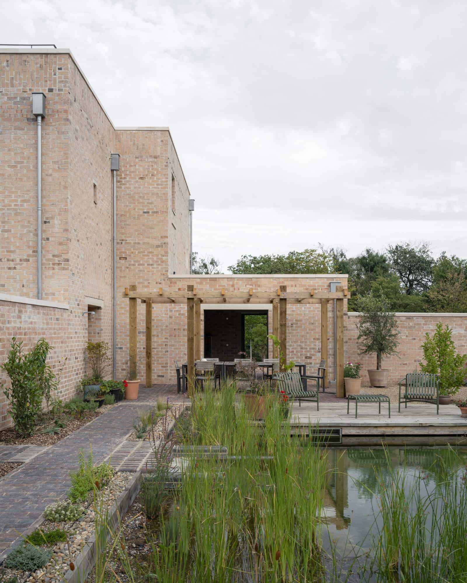 The garden courtyard of A House and Garden at Fairmead in Epping Forest, with a pale brick wall, a timber pergola and a pond edged with tall planting