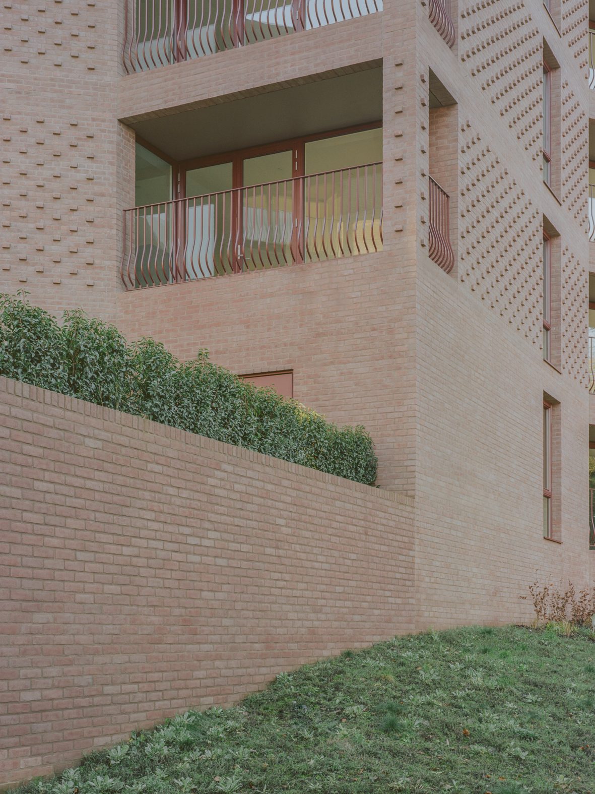 A corner of a Lion Green Road block with a cantilevered corner balcony and window, a perforated brick pattern on the walls above and a brick retaining wall topped with a hedge below