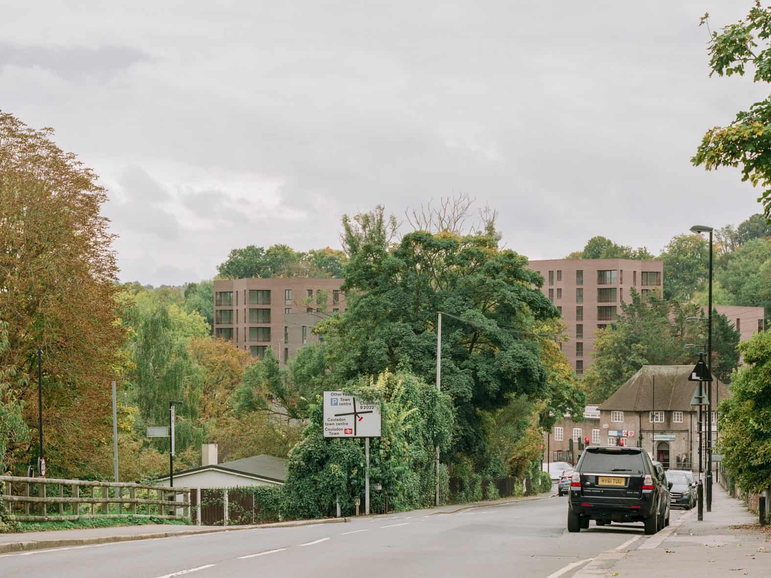 Lion Green Road in Coulsdon seen from the main road, with brown brick blocks rising behind mature trees in autumn colour beyond a road sign and passing cars