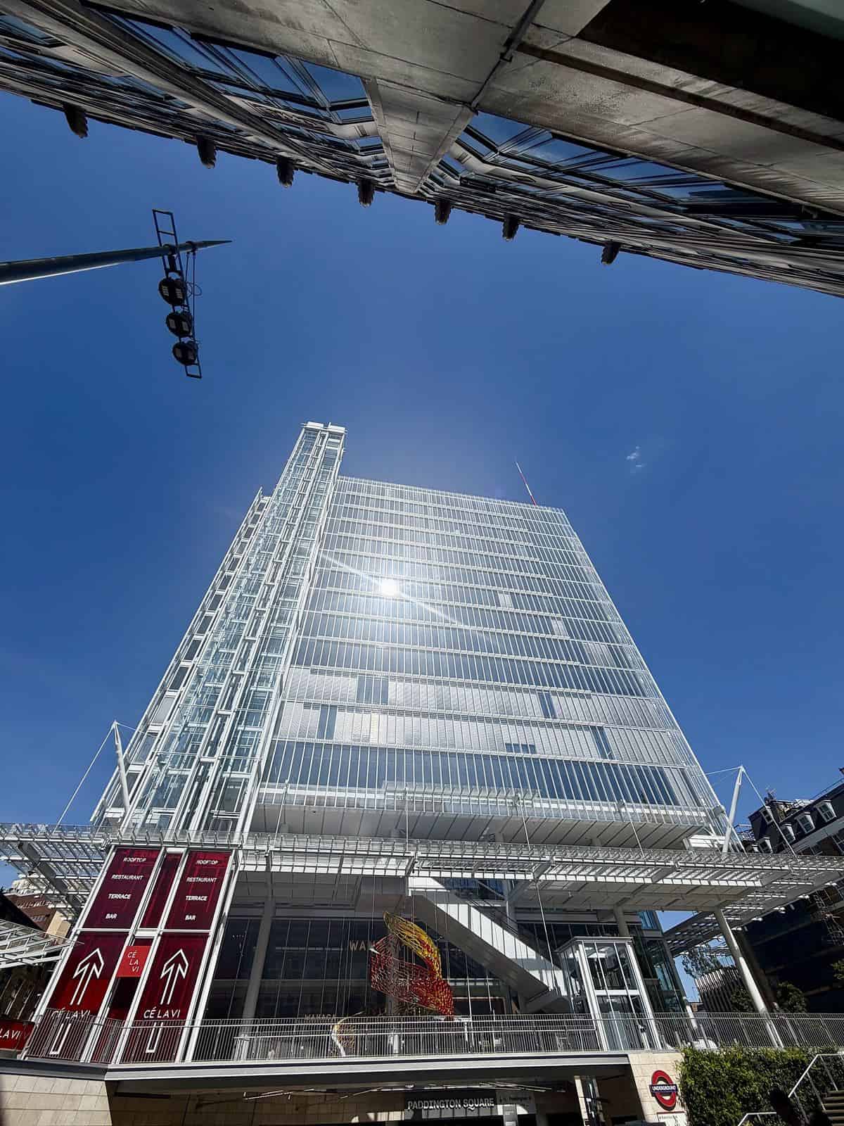 Paddington Square in London seen from below, an 18-storey glass office building with a fine steel and glass outer veil and exposed structure at its base, against a blue sky