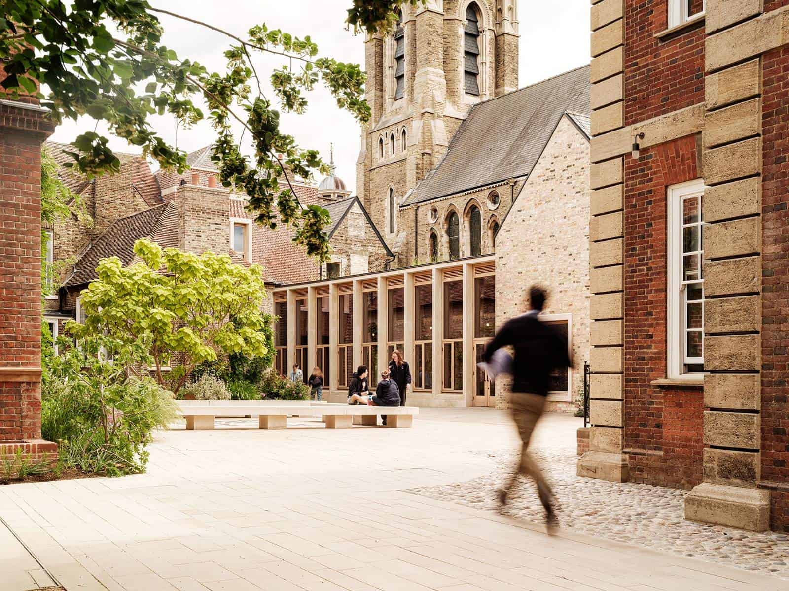 Chiu Court at Pembroke College, Mill Lane, Cambridge, a sunlit garden court framed by a red brick building, a stone church tower and trees, with a student walking across