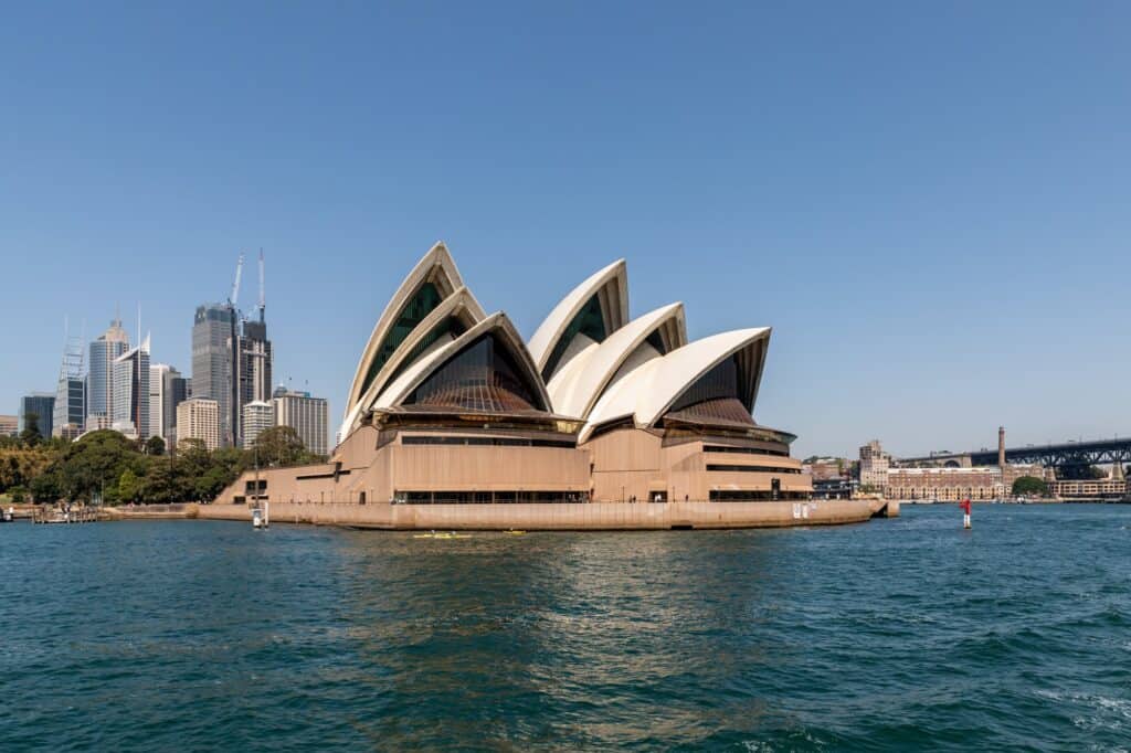 Sydney Opera House seen from the harbour on a clear day, its white tiled shells and dark glass walls rising from the pink granite podium on Bennelong Point, with the towers of the city skyline and the Harbour Bridge beyond