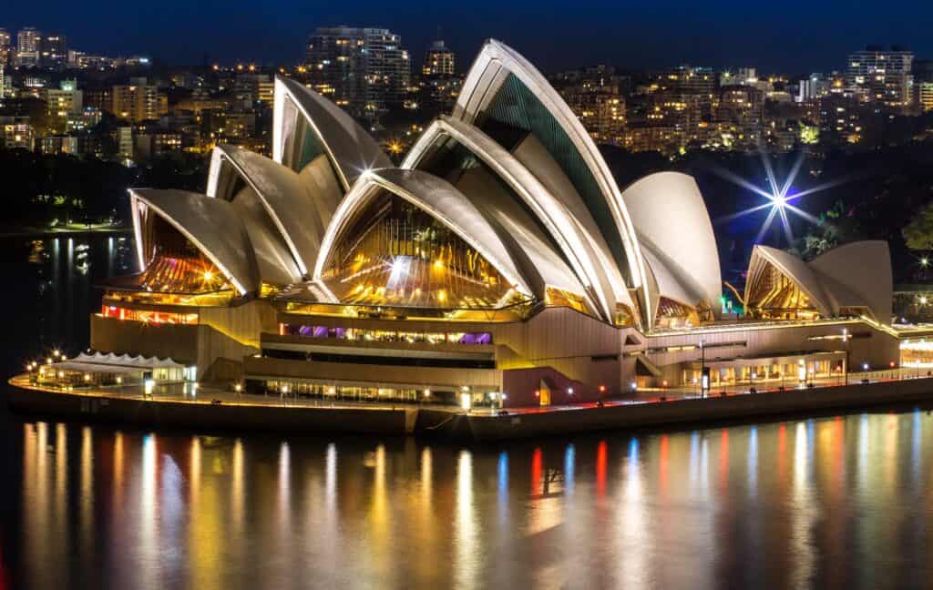Sydney Opera House lit up at night, golden light glowing through the glass walls beneath its white shells and coloured reflections rippling across the harbour, with the lights of the city hillside behind