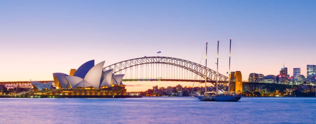 Panoramic dusk view across Sydney Harbour of the Sydney Opera House beside the Sydney Harbour Bridge, a three-masted yacht on violet water and the city skyline glowing under a pink sky