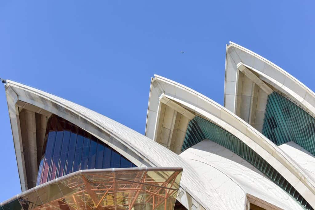 Close view of three tiled concrete shells of the Sydney Opera House against a deep blue sky, their ribbed edges framing tinted glass walls, with a bronze-framed glazed canopy in the foreground