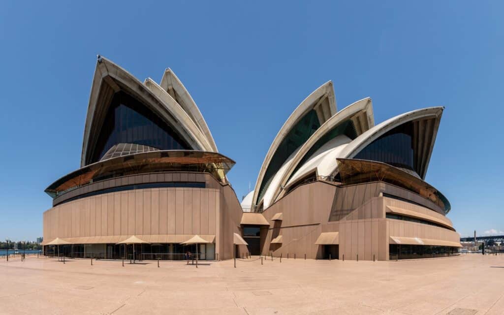 Wide view of the two main groups of shells of the Sydney Opera House rising above the curved granite-clad podium and its shaded terrace, seen across the paved concourse under a cloudless sky