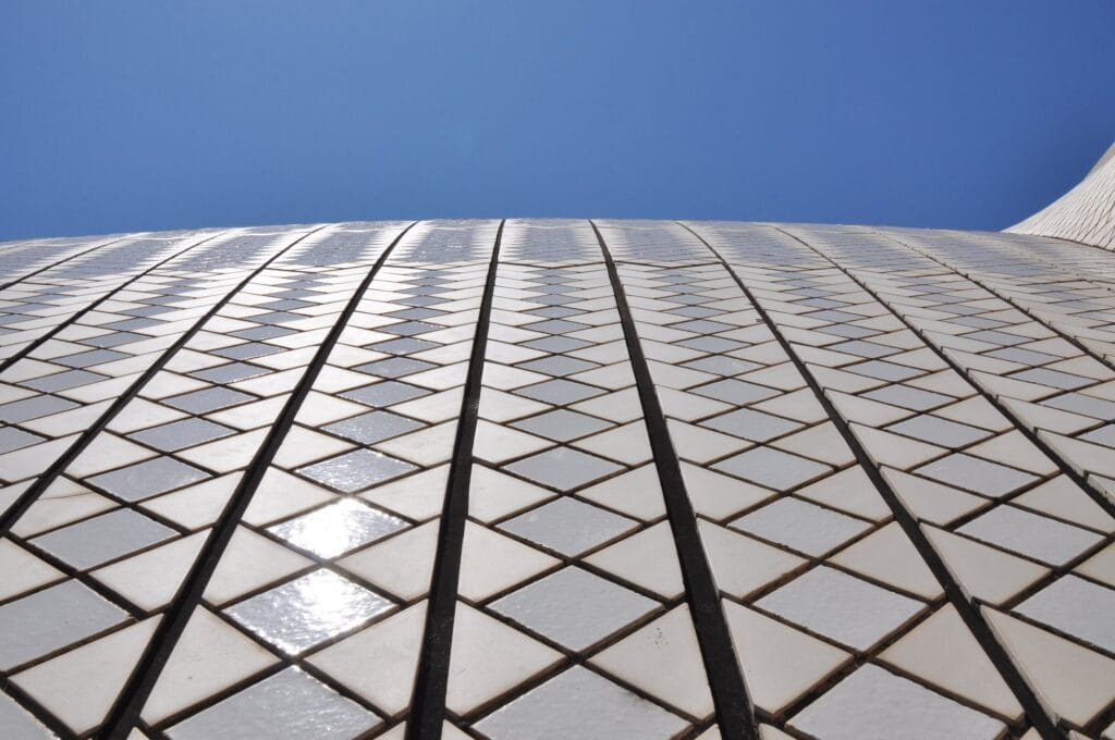 Low view along a Sydney Opera House roof showing the chevron pattern of glossy white and matte cream tiles divided by dark joints between the precast tile panels