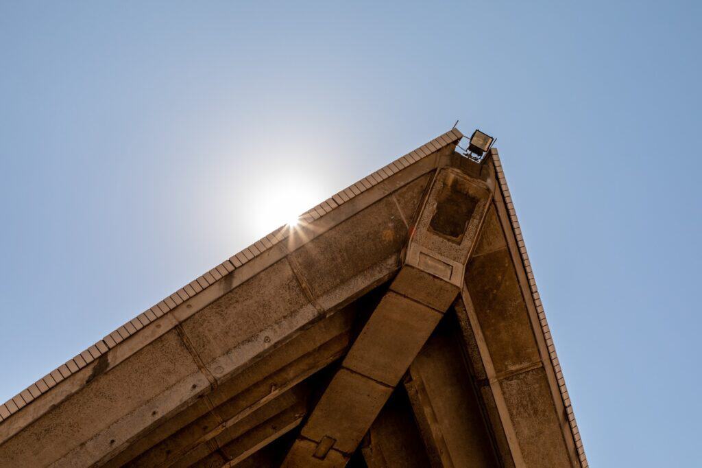 Underside of a Sydney Opera House shell tip seen from below, the exposed precast concrete ribs converging at the apex with the sun flaring behind against a pale blue sky