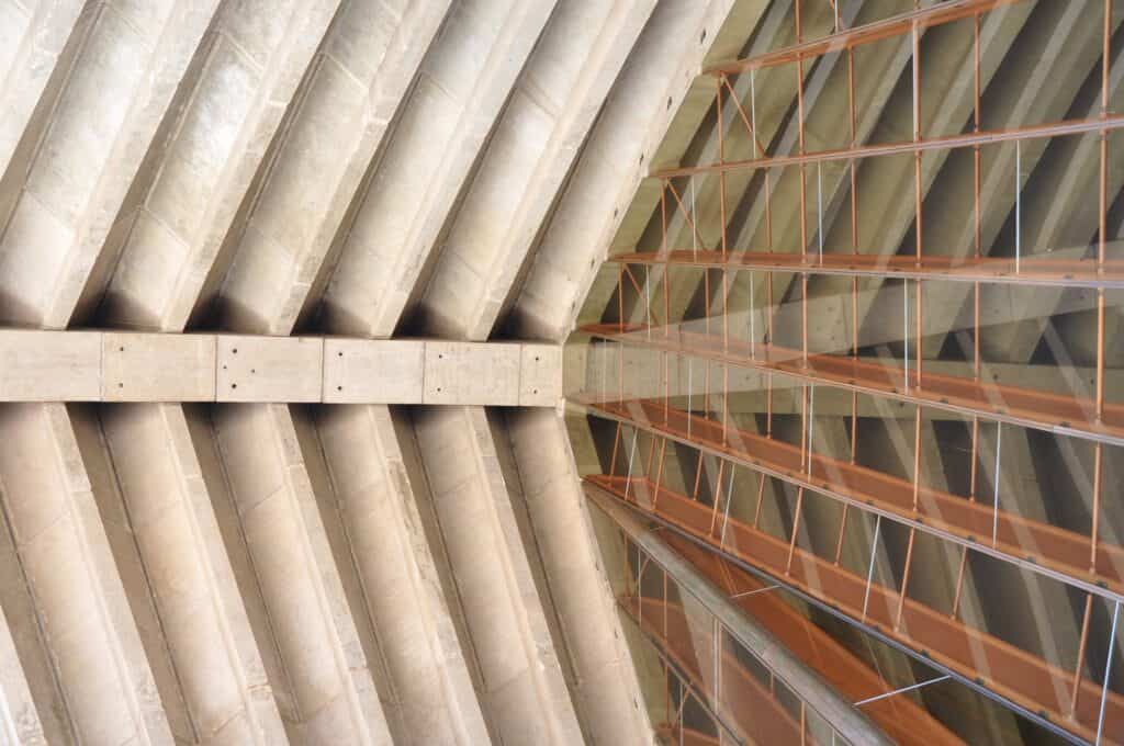 Inside the Sydney Opera House, fanned precast concrete roof ribs meeting a central concrete beam beside the great glass wall and its copper-toned steel mullions