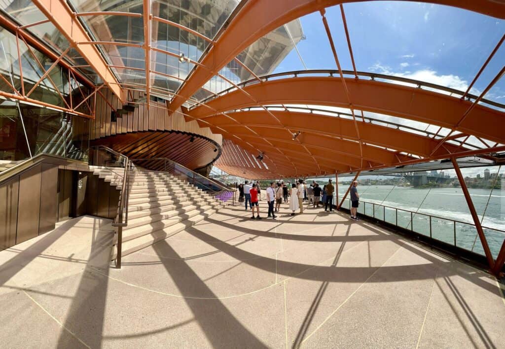 Sunlit foyer of the Sydney Opera House under curved copper-coloured steel glazing ribs, a wide stair rising beneath the timber-clad underside of the hall and visitors looking out over the harbour