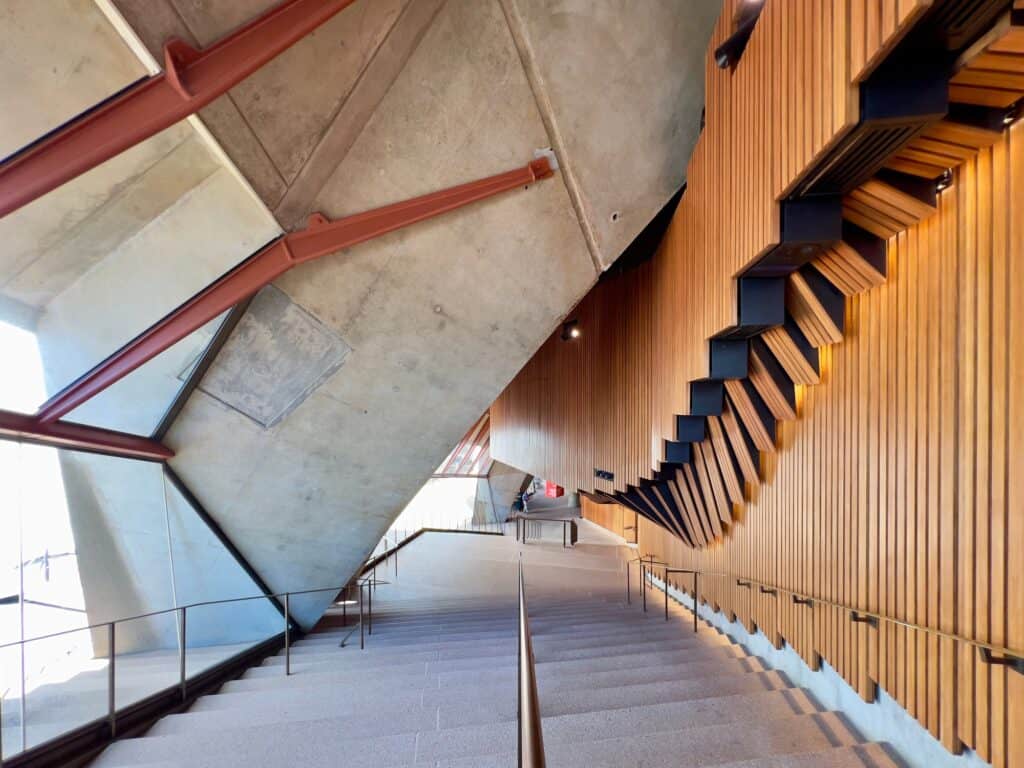 Timber-clad stepped underside of an auditorium rising above a concrete stair in a Sydney Opera House foyer, red steel struts bracing the angled concrete walls and daylight entering from the glass wall