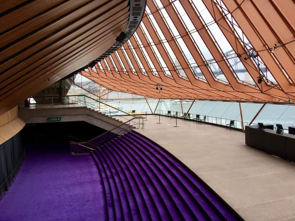 Upper foyer of the Sydney Opera House with sweeping purple-carpeted steps beneath the curved timber-ribbed ceiling and copper-toned glazing ribs, the harbour visible through the glass wall