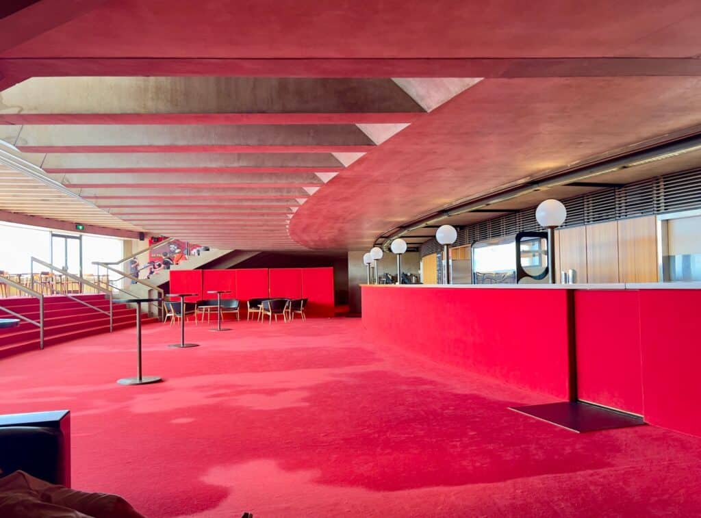Red-carpeted foyer of the Joan Sutherland Theatre at the Sydney Opera House, a red bar counter with globe lamps beneath angled concrete ceiling beams and red steps leading to harbour-view windows
