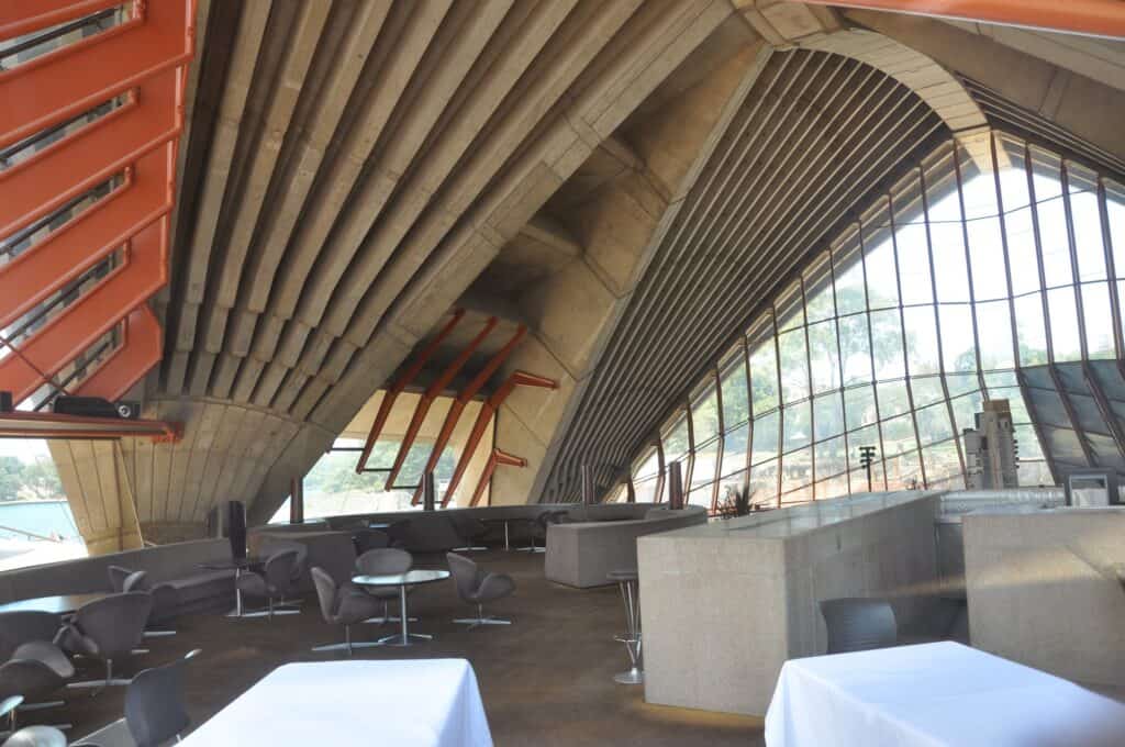 Bennelong restaurant inside the smallest group of shells at the Sydney Opera House, white-clothed tables and grey chairs beneath ribbed concrete vaults rising to a tall glass wall