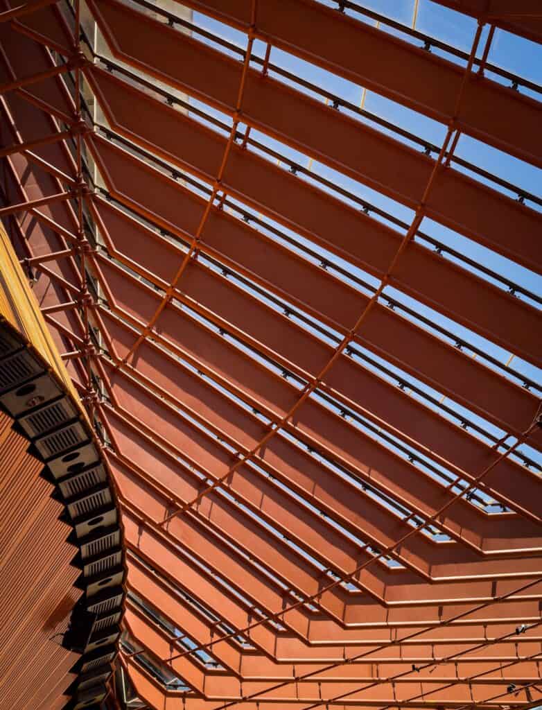 Looking up at the glass wall of a Sydney Opera House foyer, rows of copper-toned steel mullions and glazing bars framing the sky beside the timber-clad exterior of the hall