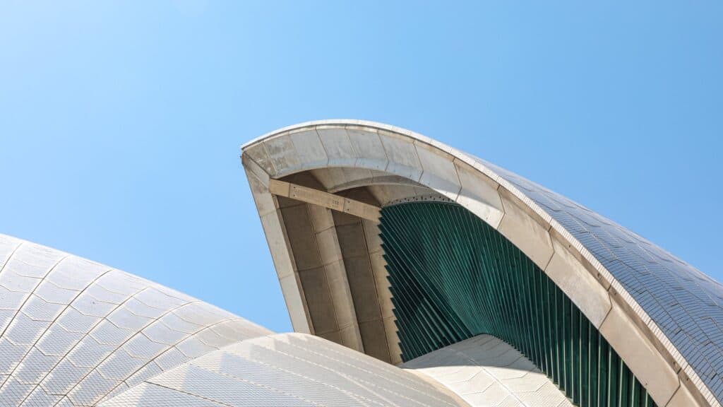 Tip of a Sydney Opera House shell against a bright blue sky, the tiled roof curving over its concrete rib edge and the green-tinted glass wall tucked beneath