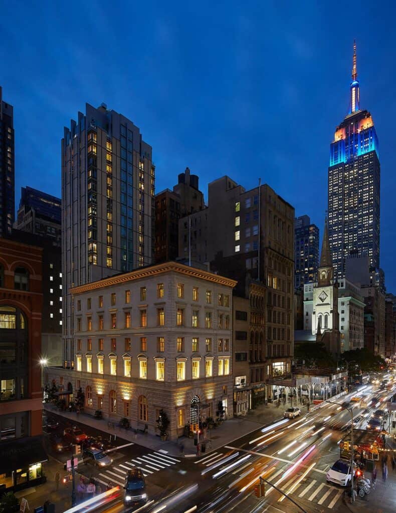 The Fifth Avenue Hotel at dusk, the lit five-storey palazzo on the corner of Fifth Avenue and 28th Street with its new tower behind and the Empire State Building beyond