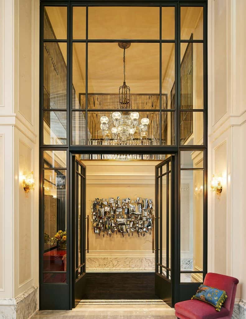 Double-height entrance of The Fifth Avenue Hotel, a tall black steel and glass screen with open doors onto a lobby with a crystal chandelier and a mirrored wall sculpture, beside a red armchair