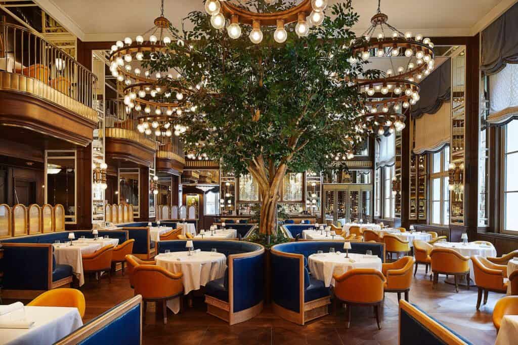 Café Carmellini in the former banking hall of The Fifth Avenue Hotel, a tree at the centre ringed by blue velvet banquettes, tiered bulb chandeliers and wood-panelled balconies