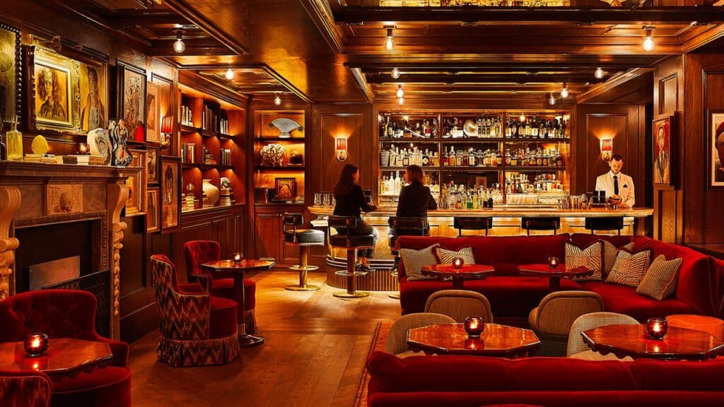 The Portrait Bar at The Fifth Avenue Hotel, a wood-panelled room hung with portraits, a carved stone fireplace, red velvet banquettes and a backlit bar