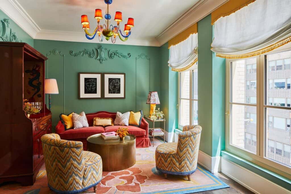 Suite living room at The Fifth Avenue Hotel with green panelled walls, a red sofa, chevron-patterned armchairs, a Murano glass chandelier and tall windows onto Fifth Avenue