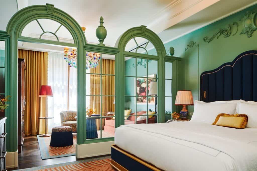 Bedroom at The Fifth Avenue Hotel divided from its sitting area by green arched glazed screens, a navy upholstered headboard and gold-curtained windows beyond