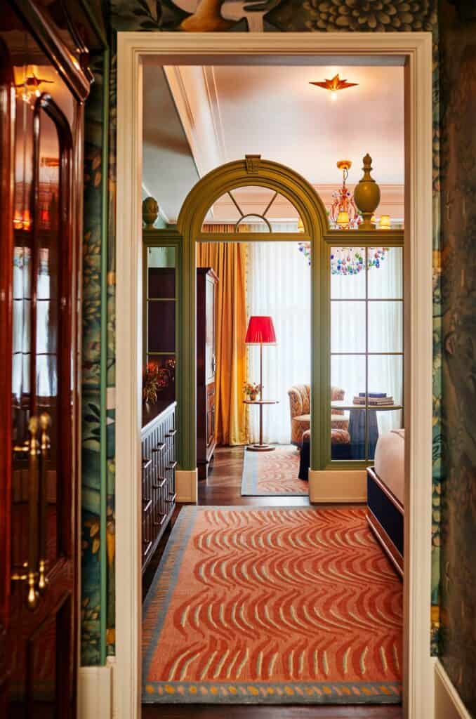 View through a doorway at The Fifth Avenue Hotel past green foliage wallpaper and a mahogany cabinet, along a wavy orange runner to a green arched screen and a red floor lamp