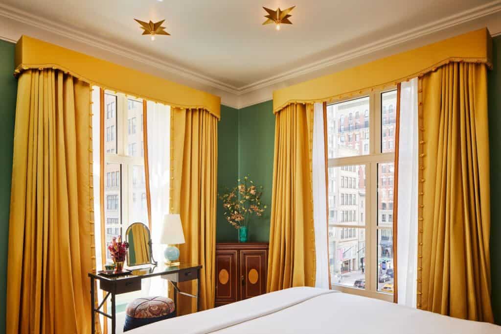 Corner bedroom at The Fifth Avenue Hotel with buttercup-yellow curtains and pelmets, green walls, star-shaped ceiling lights and a writing desk by the window