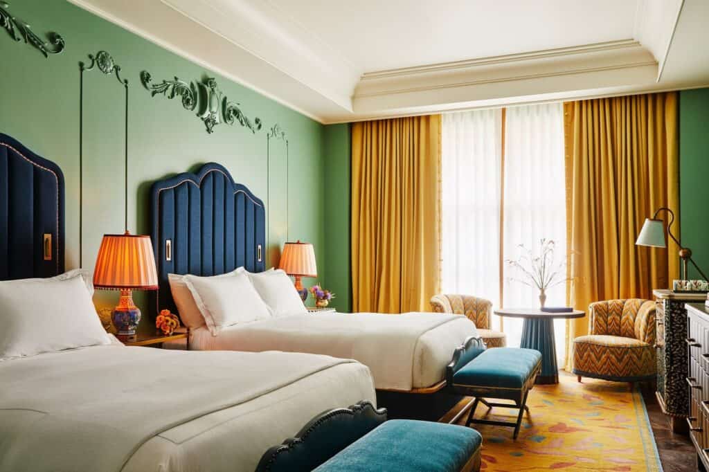 Twin room at The Fifth Avenue Hotel with tall scalloped navy headboards against green walls with plaster scrollwork, orange lamps and a yellow patterned rug