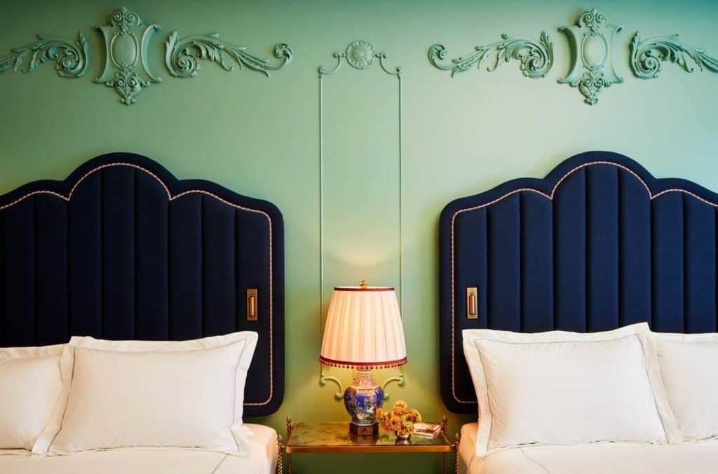 Two scalloped navy velvet headboards at The Fifth Avenue Hotel beneath green plaster cartouches, with a pleated pink lampshade on a blue and white porcelain base