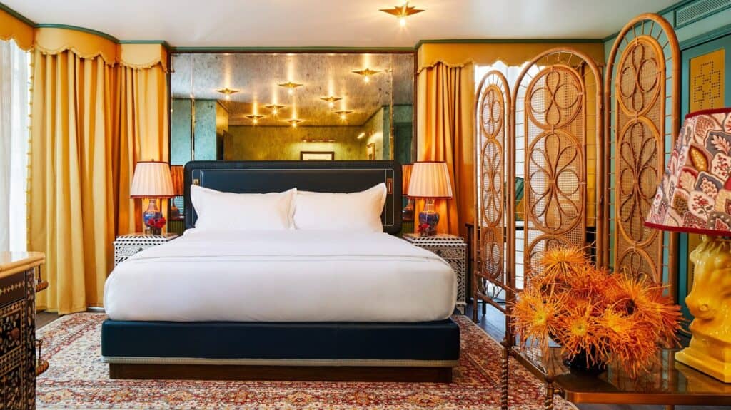 Bedroom at The Fifth Avenue Hotel with a navy upholstered bed on a Persian rug, rattan folding screens, mirrored walls and gold curtains