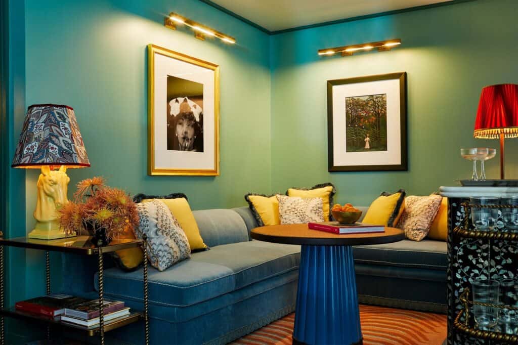 Sitting nook at The Fifth Avenue Hotel with a blue velvet banquette and yellow cushions, a fluted blue table, framed artworks under brass picture lights and an inlaid black cabinet