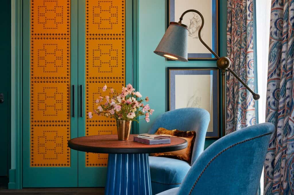 Corner of a room at The Fifth Avenue Hotel with orange studded wardrobe doors in a green frame, blue velvet armchairs, a round table with pink flowers and an angled floor lamp
