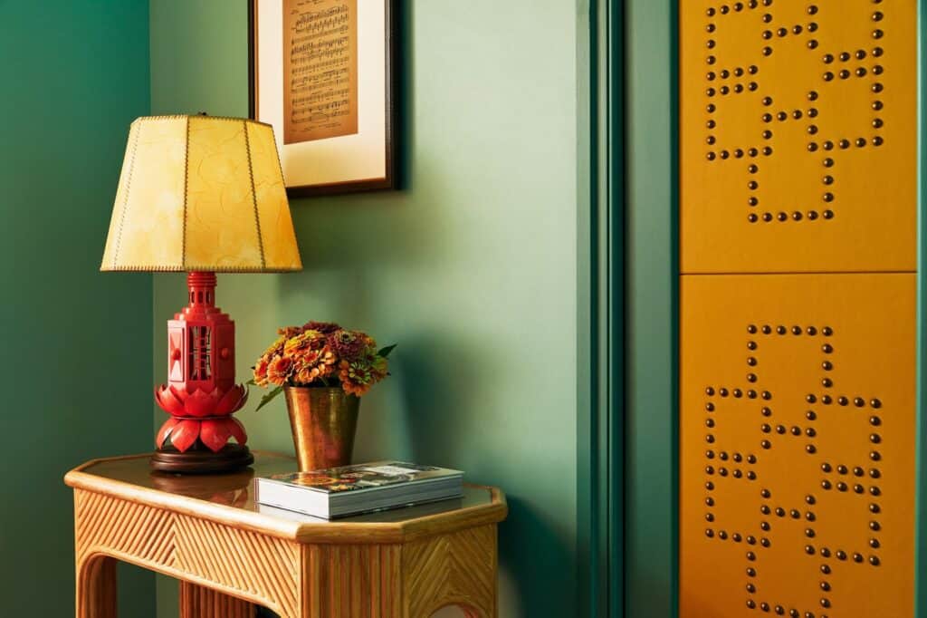 Bedside detail at The Fifth Avenue Hotel, a red lacquered lamp with a parchment shade on a rattan console beside green walls and a studded mustard panel