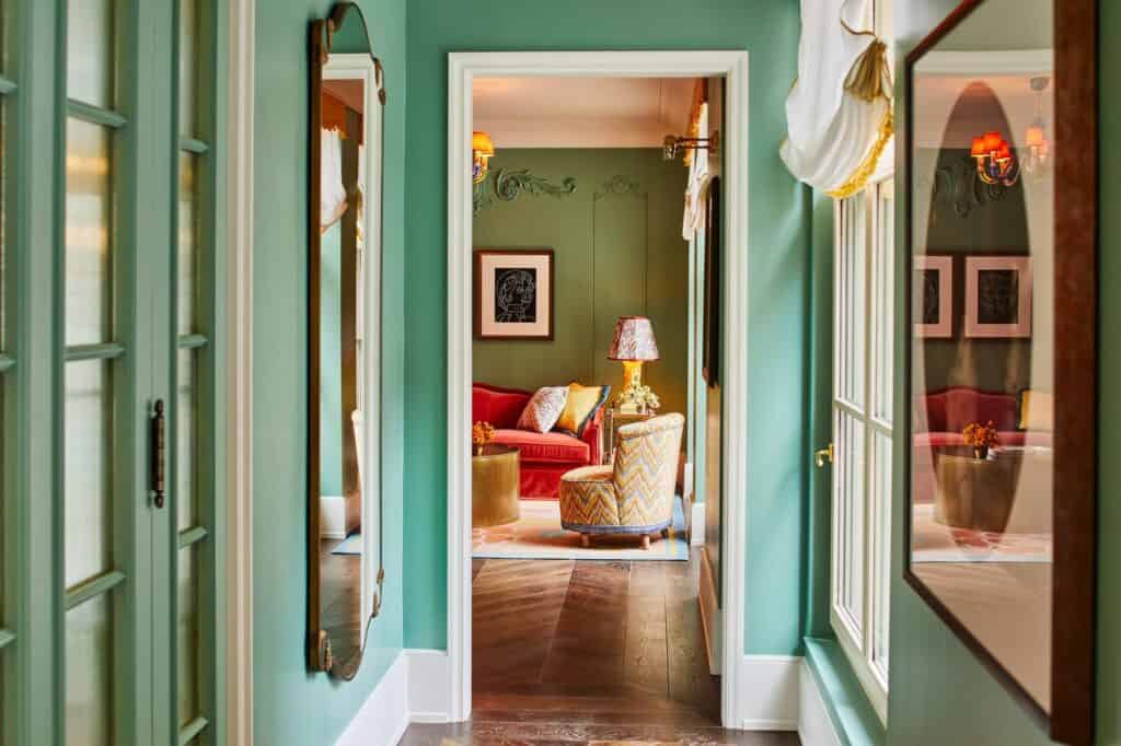 Green-painted hallway of a suite at The Fifth Avenue Hotel with glazed doors and oval mirrors, opening onto a living room with a red sofa and a patterned armchair