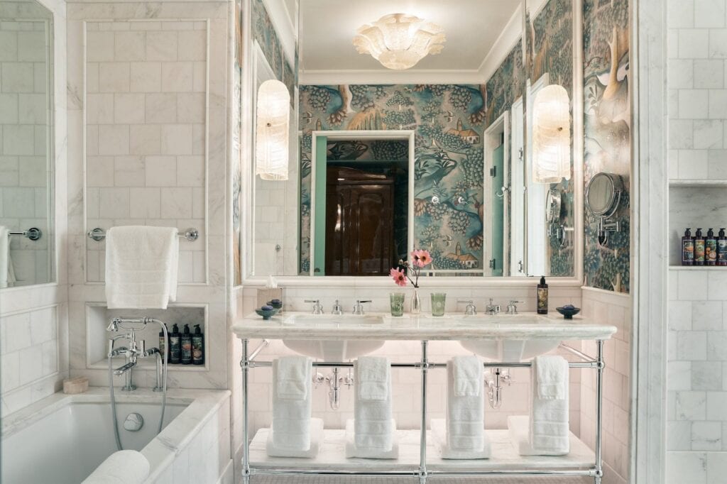 Marble bathroom at The Fifth Avenue Hotel with a double washstand on nickel legs, blue-green bird and foliage wallpaper, a shell-shaped ceiling light and a white-tiled bath