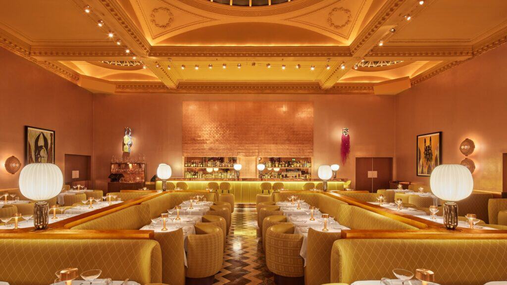 Axial view down the Gallery at Sketch towards the glowing yellow bar, sunshine-yellow banquettes and white-clothed tables lit by white globe lamps, copper-papered walls hung with Yinka Shonibare artworks beneath a gilded ornate ceiling