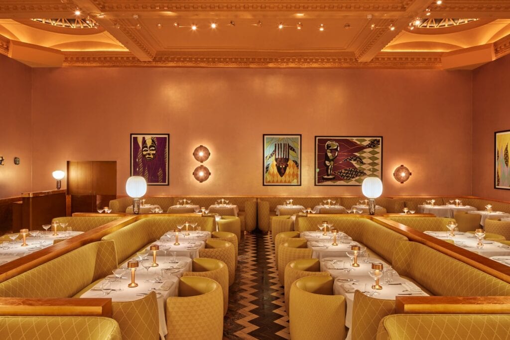 Symmetrical view across the Gallery at Sketch, rows of tables between quilted yellow banquettes and tub chairs either side of a chevron-patterned aisle, three Shonibare quilts and paired round wall lights on the copper end wall