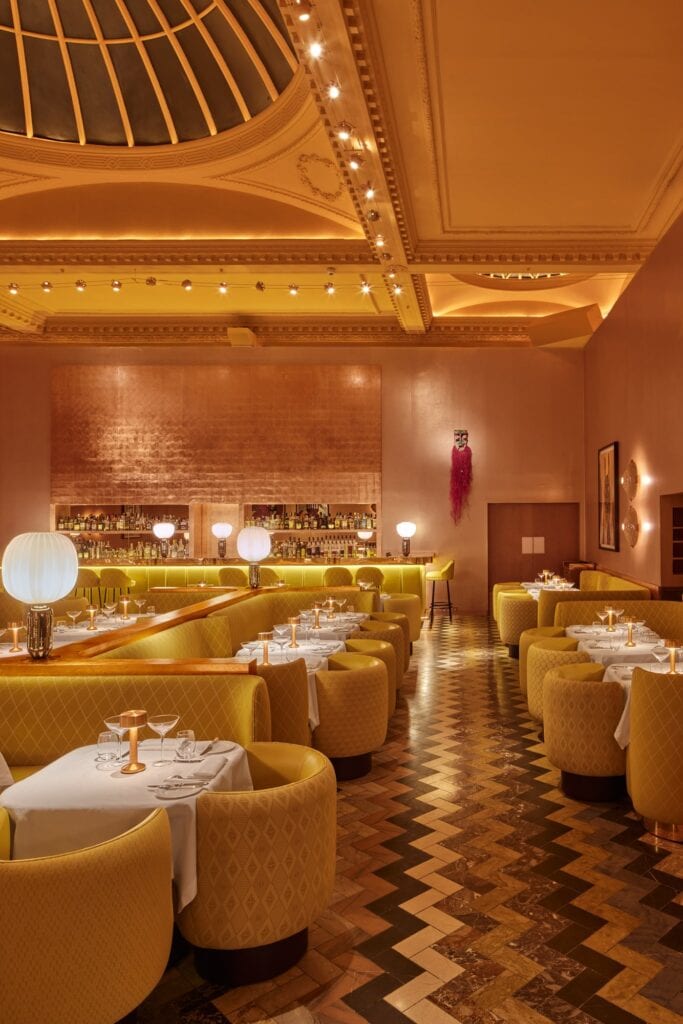 Tall view of the Gallery at Sketch beneath its glazed lantern skylight, a herringbone floor leading between yellow tub chairs and tables to the bar, with a white globe lamp and a pink raffia mask on the copper wall