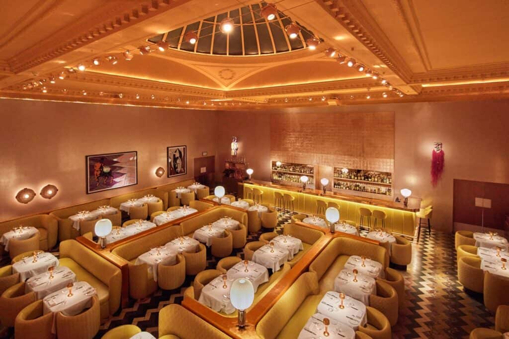 Elevated view over the Gallery at Sketch with the domed skylight overhead, islands of yellow banquettes and white tables on a dark and light chevron floor, framed artworks and the golden-lit bar beyond