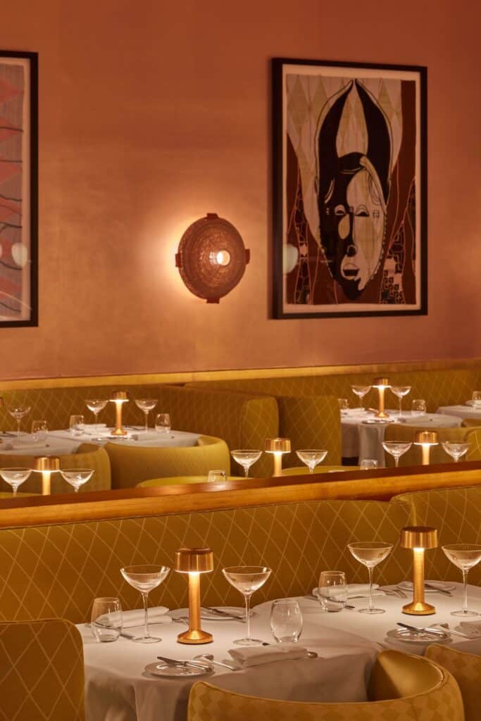 Framed Yinka Shonibare quilt of a stylised mask above a quilted yellow banquette at the Gallery at Sketch, beside a round woven wall light, with brass table lamps and coupe glasses on the white tables
