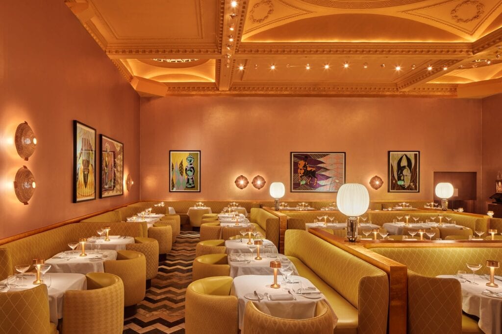 Angled view of the Gallery at Sketch, pairs of round woven wall lights and framed Shonibare quilts on the copper walls above long yellow banquettes, globe lamps and set tables on a chevron floor