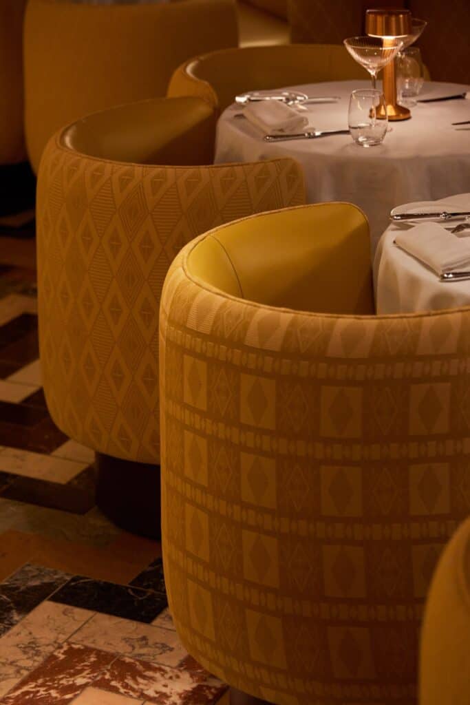 Close view of curved tub chairs upholstered in textured yellow raffia fabric at the Gallery at Sketch, set around a white-clothed table with a brass lamp and glassware