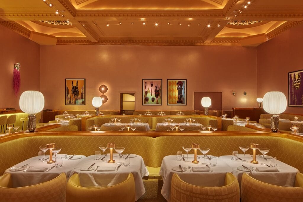 Banquette tables at the Gallery at Sketch laid with white cloths, coupe glasses and brass lamps in front of quilted yellow upholstery, large white globe lamps and three framed Shonibare artworks beyond