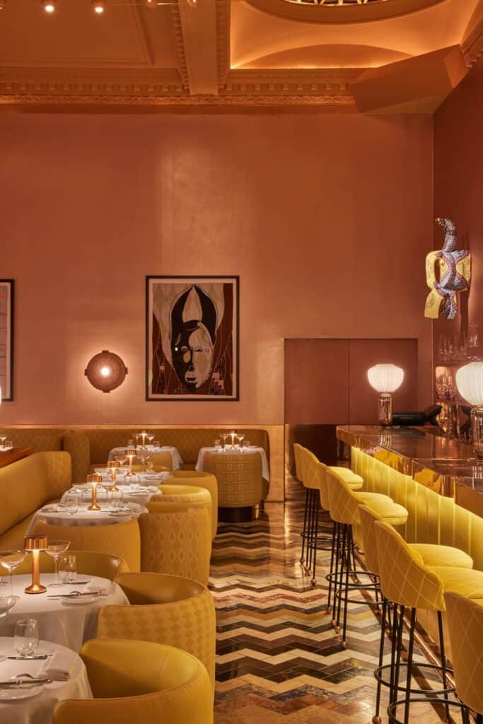 Bar at the Gallery at Sketch with yellow-upholstered stools along a gold counter, a carved figure sculpture, a pink raffia mask on the copper wall and a framed mask quilt above the dining room